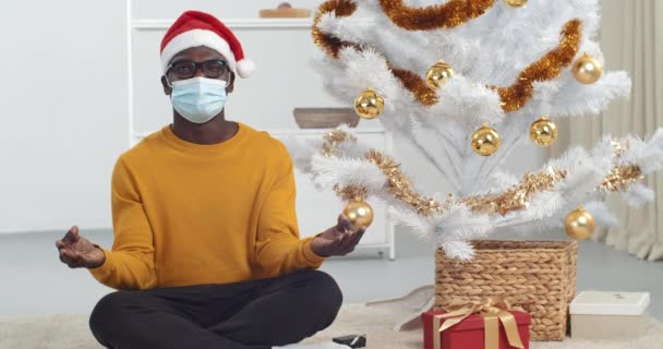 Afro americano cara preto homem sentado no chão sob árvore de natal em posição de lótus praticando exercícios respiratórios meditando estresse luta para o feriado de ano novo ouve barulho abre um olho olha ao redor — Vídeo de Stock