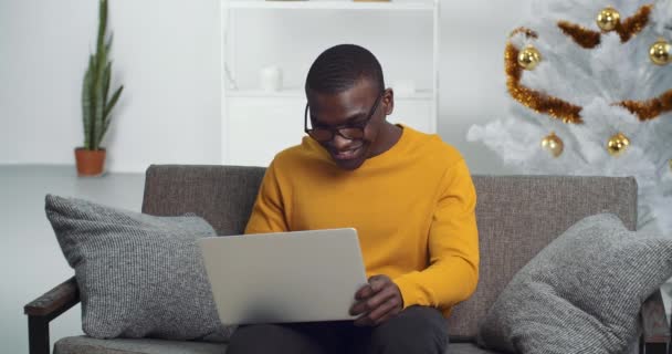 Porträtt av afro amerikansk koncentrerad affärsman sitter på soffan i vardagsrummet nära julgran håller bärbar dator skriva studera i helgdagar arbetar på distans hemifrån för nytt år på grund av tidsfrister — Stockvideo