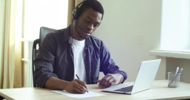 Handsome afro american man black guy in headphones student teacher speaks online remotely via video call chat conference studying remotely writes notes with pen on paper, work study during quarantine