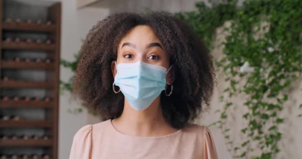 Tıbbi koruyucu maskeli Afro-Amerikan kız portresi hemşire öğrenci, kıvırcık saçlı siyah kadın elini sallıyor ve kameraya merhaba diyor ve uzaktan konuşuyor. — Stok video