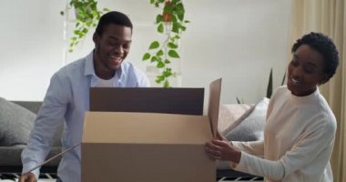Afro-Amerikan çift karı koca karton kutuyu açarlar. Karton kutuya bakarlar. Paketin içindeki küçük kız mutlu bir şekilde güler.