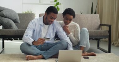 Young black man holding credit banking card payment information with focused smiled millennial afro american woman doing online shopping order, happy couple involved in purchasing goods in internet