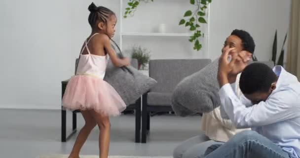 Афро-американская семья мама папа и девочка ребенок в розовом платье, как балерина борется с серыми подушками сидя на полу в гостиной дома весело играть вместе смеясь, родительские концепции любви — стоковое видео