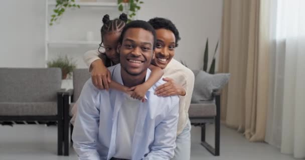 Home portrait, happy afro american woman and baby girl hug smiling black man by neck. Loving young seven, wife mom and daughter hug dad husband from behind looking at camera sincere facial expressions — Wideo stockowe