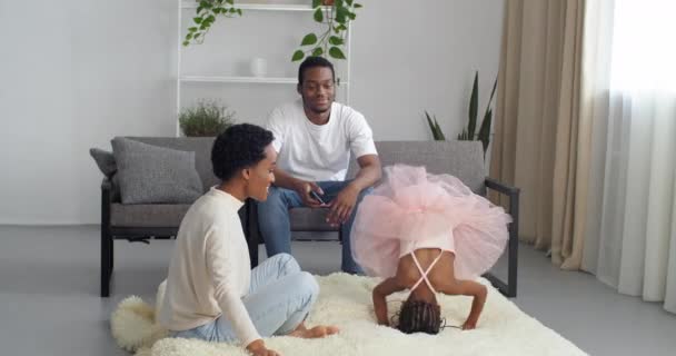 Junge Eltern afroamerikanische Familie verbringen Zeit mit niedlichen aktiven verspielten Tochter in rosa Kleid sitzt auf dem Sofa zu Hause im Wohnzimmer beobachten Mädchen Kind springen Vater macht Foto auf Handy — Stockvideo