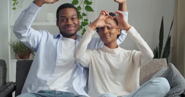 Portrait of afro american couple girlfriend and boyfriend sitting together on sofa at home in living room showing heart shape sign of love symbol of sympathy valentine, concept of happy relationship — Wideo stockowe