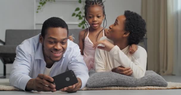 Afro american ethnic family taking photo on mobile phone camera posing having fun lying together looking at screen of smart phone three people spending time at home interior, love and support concept — Stockvideo