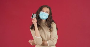 Studio portrait caucasian girl melinial masked woman holding credit card in her hand pensive comes up with idea solution rejoices jumping with happiness, concept of remote order winning lottery money