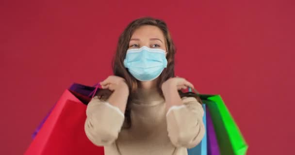 Lycklig kvinna i medicinsk mask skyddar ansiktet från infektion med respiratoriskt virus håller färgglada shoppingpåsar i händerna glädjer shopping efter gåvor nya kläder shopping koncept under pandemi — Stockvideo