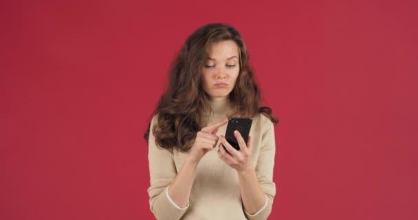 Portrét kavkazské ženy s dlouhými vlasy stojí na červeném pozadí v ateliéru drží telefon v ruce surfování Internet přichází s nápadem vytáhne bankovní kreditní karty dělá nákup on-line obchod koncept — Stock video