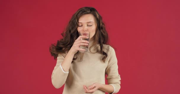 Studio shoot jeune femme avec de longs beaux cheveux se tient sur fond rouge en chandail blanc tient le verre dans sa main boit propre savoureuse eau minérale froide, se sent déshydratation soif, concept de soins de santé — Video