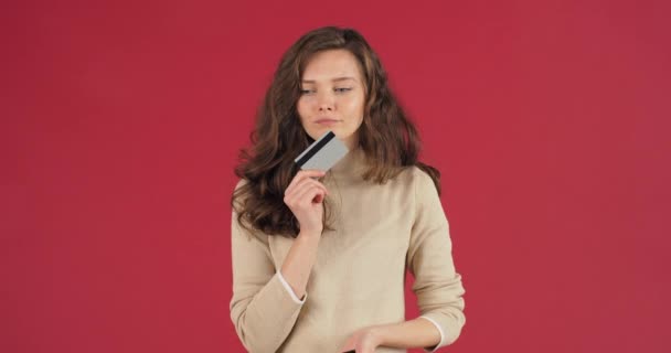 Young beautiful girl on red background posing with bank credit card in her hand comes up with idea for gift orders buys online through smartphone mobile phone enjoys discounts spends money ecommerce — Stock Video