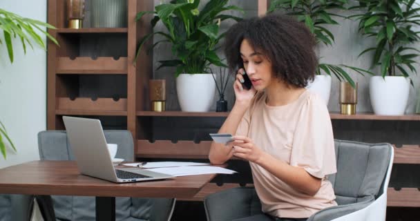 Young zaměřený afro americká etnická žena sedí v kavárně restaurace nakupování on-line s kreditní kartou mluvení mobilní telefon žena pomocí notebooku nákup objednávky doručení v internetovém obchodě — Stock video