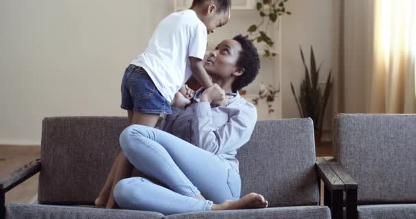 Afro american mom with daughter on couch hugging tenderly cuddling communicates talking joking smiling spending time together at home on weekend, motherhood and parenthood concept, ethnic family love — Stok Video