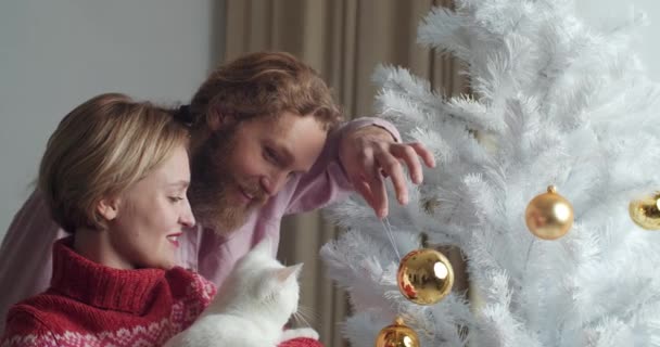 Husband and wife decorate Christmas tree, woman holds white cat in her arms, man plays with pet yellow gold Christmas ball, caucasian couple family prepares for home winter holidays together close-up — ストック動画