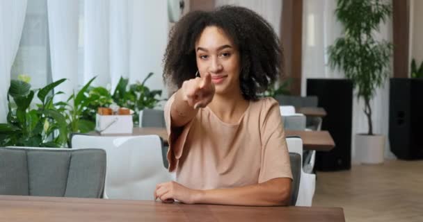 Portrait belle frisée mixte race fille adolescente femme assise à la table dans le café en regardant la caméra pointe doigt devant elle-même invite à son petit ami garçon homme à venir faire geste de la main — Video