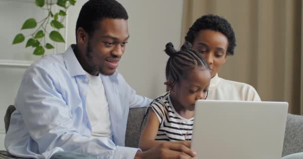 Молодые афро-американские родители мама и папа семья научить маленькую милую дочь девочка использовать ноутбук Интернет-компьютер для обучения развития сидя на диване дома, родительство и счастливое детство концепции — стоковое видео