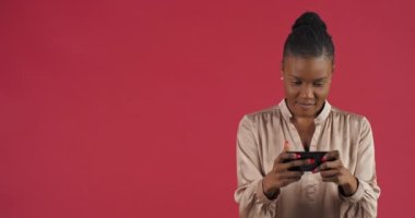 Neşeli, konsantre Afro-Amerikan kadın etnik milenyum kızı Red Stüdyo arka planında duran akıllı telefon zımbırtısıyla mobil oyun oynayan genç mutluluğun zaferini kutluyor.