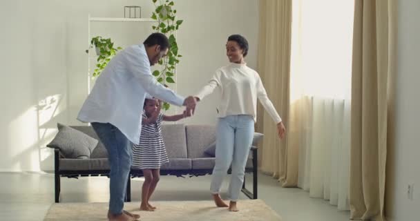 Etnisk afrikansk amerikansk familj unga par mamma och far förälder tillbringa tid med flicka barn dansa med dotter i vardagsrummet hålla händer njuta av roliga part skrattar leende fullängdare filma — Stockvideo