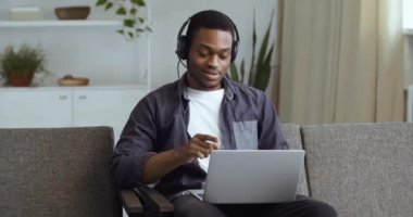 Siyah adam Afro Amerikalı işadamı, kulaklıklı genç öğrenci, dizüstü bilgisayarla müzik dinliyor, videolar dinliyor, şarkı söylüyor, dans figürleri yapıyor, evdeki kanepede dinleniyor.