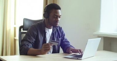 Afro-Amerikalı adam internetten kulaklıkla müzik dinliyor elinde plastik banka kredi kartı var ağda internetten alışveriş yapıyor ev masasında oturarak indirimli dans kutluyor. 