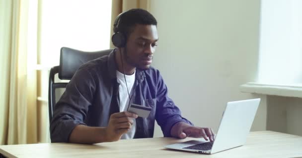 Afro amerykański facet słucha muzyki online ze słuchawkami trzyma plastikową kartę kredytową banku w ręce sprawia, że zakup zamówienia online w sieci świętuje szczęśliwe zniżki taniec siedzi przy stole w domu — Wideo stockowe