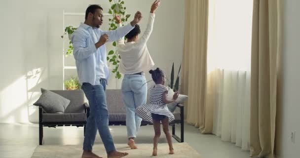 Αφρο-αμερικανική γυναίκα μαύρος άνδρας και μικρή εθνοτική κορίτσι που διασκεδάζουν μαζί στο σαλόνι κοντά στον καναπέ στο εσωτερικό του σπιτιού περνούν χρόνο μαζί χορεύοντας κινήσεις του σώματος στη μουσική, νεαρή οικογένεια τρία άτομα — Αρχείο Βίντεο