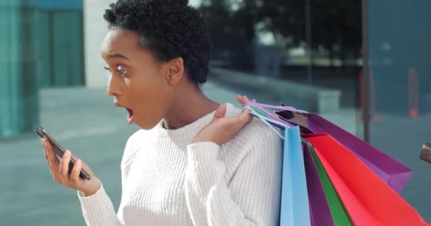 Afro americká žena smíšené rasy shopaholic dívka nakupující spotřebitel se podívá na telefon dostane emeil s fakturou z obchodu cítí šok překvapen vysokými náklady na nové oblečení a dárky drží tašky nákupů