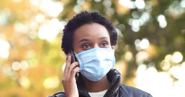 Portret afro amerykańskiej kobiety w masce medycznej na twarzy używa telefonu komórkowego do zdalnej komunikacji podczas pandemii na zewnątrz jesienią, zagraniczny student freelancer mówi przez smartfona online — Wideo stockowe