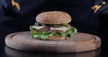 Ağır çekimde yakın plan kesilmiş siyah eldivenli tohum erkek elleri ahşap tahtanın üzerine patates kızartması koyuyor. Yürekli, iştah açıcı burgerin yanına. Erkek aşçı, bar restoranında servis edilen fast food servisini hazırlıyor.