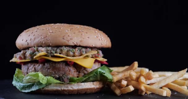Rotating shot van smakelijke dubbele Burger met vlees rundvlees salade tomaten kaas en uien staat op zwarte tafel hamburger met friet en chips tegen donkere achtergrond close-up, Amerikaanse traditie voedsel — Stockvideo