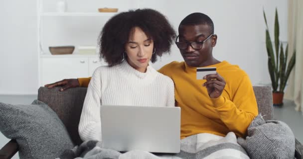 Joven pareja afroamericana hace la compra en línea a través de la tienda de Internet utilizando el ordenador portátil, ingrese el código secreto de la tarjeta bancaria de plástico, solicite un préstamo, ordene alimentos de forma remota desde casa durante la cuarentena epidémica — Vídeo de stock