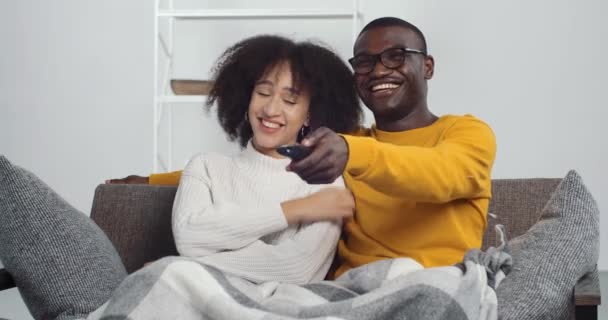 Młoda, wesoła afrykańska para siedząca na kanapie w salonie i oglądająca program do kina komediowego w telewizji śmiejąca się razem. etyczne kręcone kobieta dziewczyna i czarny mężczyzna relaks — Wideo stockowe