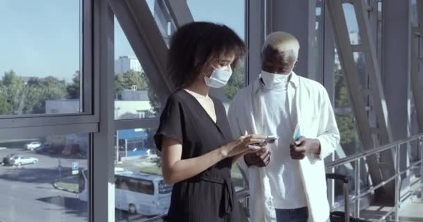 Afro american couple young friends curly girl and black handsome guy wearing protective medical masks on faces standing at airport together looking at mobile smartphone looking for flight information — Stock Video