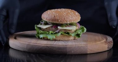 Erkek eli eldivenli yakın çekim ahşap bir tahtanın üzerindeki masadan iştah açıcı kokulu yağlı hamburgeri alır. Salata peynirli Chisburger siyah arka planda duruyor, sağlıksız fast food konsepti.
