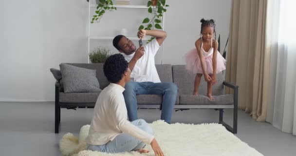 Τρεις άνθρωποι νέοι αφρο-αμερικανική οικογένεια κάθεται στο σαλόνι εσωτερικό του πατέρα παίρνει τηλέφωνο φωτογραφική μηχανή παίρνει φωτογραφία, ενώ η μικρή κόρη πηδά από καναπέ σε πάτωμα στο χαλί δίνει μαμά πέντε σύμβολο της υποστήριξης — Αρχείο Βίντεο