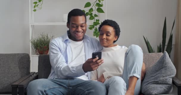 Retrato de homem afro-americano namorado e mulher étnica namoradas sentadas juntas no sofá abraçando olhando para a tela do telefone do gadget móvel smartphone moderno conversando com amigos recebem mensagem — Vídeo de Stock