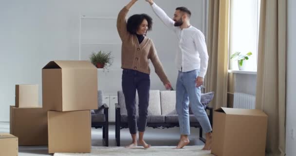 多民族夫妻手牵着手在客厅里的纸板箱旁跳舞，庆祝着搬到新家去换公寓，快乐的家庭未来理念 — 图库视频影像