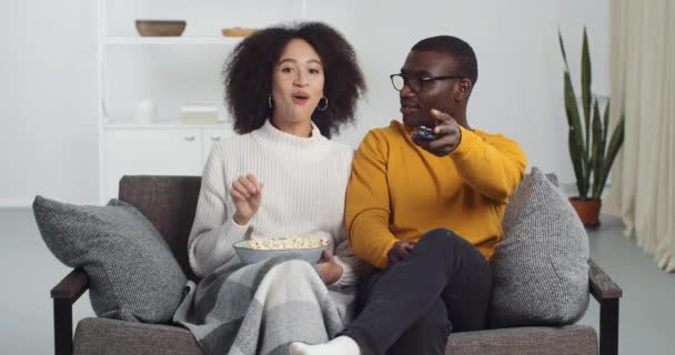 Afro amerikansk familj par lockig kvinna och man i glasögon sitter tillsammans på soffan titta på tv komedi film äta popcorn skratta ha kul i vardagsrummet, byter kille kanaler med fjärrkontroll — Stockvideo