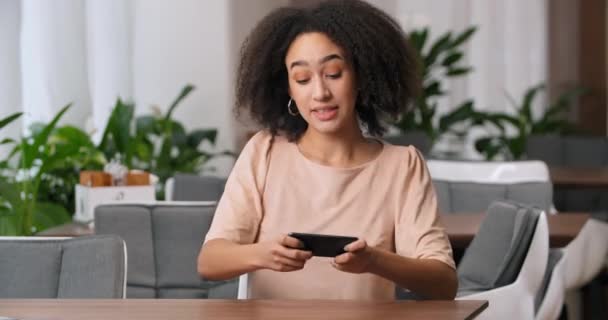 Brincalhão alegre concentrada africana americana mulher étnica menina milenar adolescente jogando jogo móvel no gadget smartphone ganha regozija vitória triunfo levanta as mãos com alegria — Vídeo de Stock