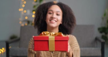 Mutlu cömert afro-amerikalı kadın kameraya büyük kırmızı hediye kutusunu gösteriyor altın kurdeleli hediyesini uzatıyor evdeki kanepede oturuyor ışıkların arka planında oturuyor
