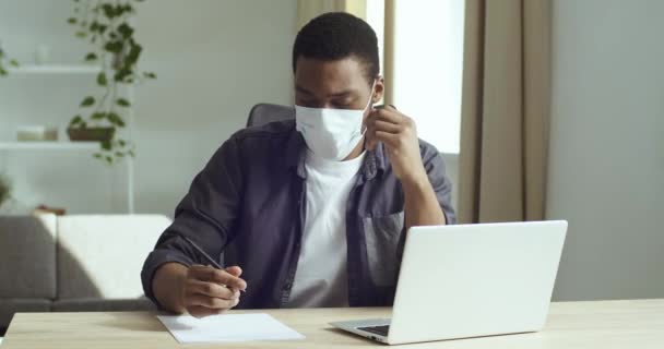 Retrato afro americano hombre de negocios estudiante en máscara médica escribe en la lista de planes de contrato de papel hace el trabajo de papel se sienta en la mesa delante de los trabajos portátiles durante la cuarentena coronavirus, concepto de infección — Vídeos de Stock