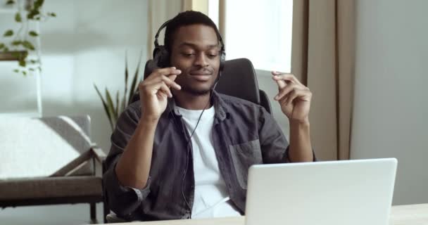 Ontspannen Afro-Amerikaanse mannelijke freelance student met casual shirt en koptelefoon aan tafel op kantoor luisteren naar muziek genieten van rust sluit zijn ogen in plezier beweegt zijn handen te verslaan van lied — Stockvideo