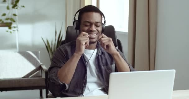 Un homme d'affaires africain assis à table sur son lieu de travail devant un ordinateur portable porte des écouteurs écoutant de la musique fait semblant de jouer de la batterie bouge les mains comme un musicien aime la chanson reposant sur la pause à la maison — Video