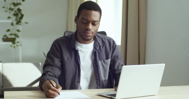Jonge man student duur afro-Amerikaanse zakenman freelancer zit aan tafel in de voorkant van laptop werken studies schrijft met pen op wit papier in document plannen analyseert doet papierwerk in het thuiskantoor — Stockvideo