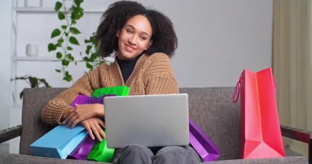 Mulher afro-americana adolescente com laptop senta-se no sofá sofá no interior da sala de estar abraços sacos de compras sacos coloridos apreciando pacotes de presentes, conceito de compras on-line descontos venda — Vídeo de Stock