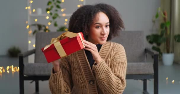 Afro americano adolescente cumpleaños chica recibe paquete de un mensajero presente entrega a domicilio sostiene caja de regalo rojo sacude unpacks siente interés curiosidad concepto de cumpleaños sorpresa vacaciones en sala de estar — Vídeos de Stock