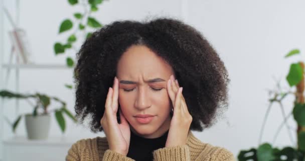 Retrato de una mujer afro-americana de pelo rizado y triste cansada que siente dolor de cabeza fiebre que sufre de un síntoma de migraña supera el estrés cogida de la mano detrás de la cabeza pensando en el plan de ideas recuerda información — Vídeos de Stock
