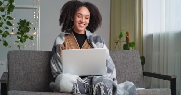 Afroamerikanische Studentin sitzt zu Hause Wohnzimmer auf Couch, mit Decke bedeckt hält Laptop Antworten Videoanruf spricht Fernbedienung auf Online-Konferenzwellen Gruß an Webcam, Quarantäne-Kommunikation