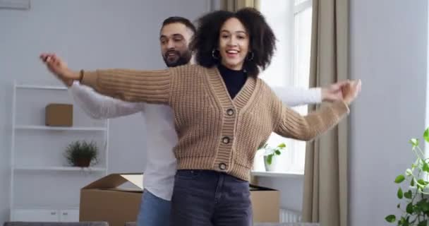 Enthousiaste jonge volwassene twee mensen, familie portret Afro-Amerikaanse vrouw vrouw en Kaukasische man man hand in hand verplaatsen naar muziek dansen tijd doorbrengen samen in nieuw huis vieren goed nieuws — Stockvideo